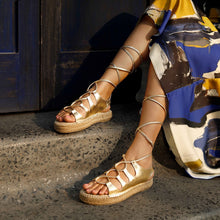 Load image into Gallery viewer, A girl sitting near a blue door on stairs wearing a pair of gold Gladiator Sandals, featuring metallic gold straps that wrap around the ankle and lower calf.