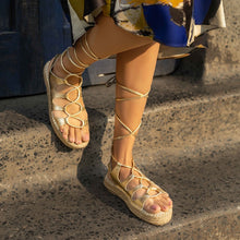 Load image into Gallery viewer, A girl walking down through stairs wearing a pair of gold Gladiator Sandals, featuring metallic gold straps that wrap around the ankle and lower calf.