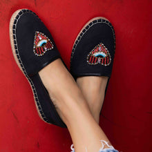 Load image into Gallery viewer, Cross legged feet of a model wearing black coloured Sweetheart Espadrilles ladies shoes with an evil eye protector design standing on a red surface