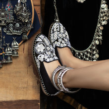 Load image into Gallery viewer, A foot of lady who have worn a Nomad Espadrilles Black shoe for women kept upon some wood with some metal jewelry in the background.
