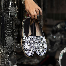Load image into Gallery viewer, A woman holding a pair of Nomad Espadrilles Black shoes for women with a metal jewelry in the background.
