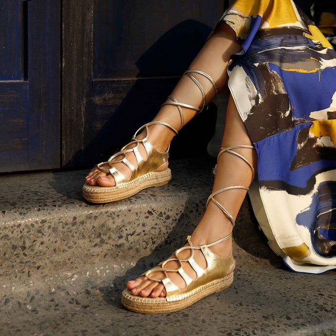 A girl sitting near a blue door on stairs wearing a pair of gold Gladiator Sandals, featuring metallic gold straps that wrap around the ankle and lower calf.