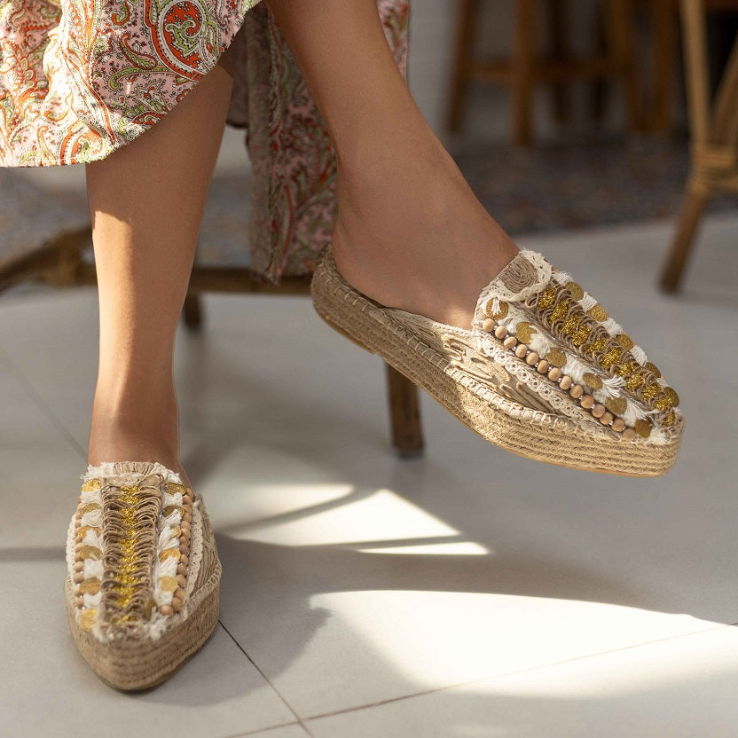 a woman's feet in golden Espadrilles on a white floor with a small detailing on them with a pattern around the bottom and sides