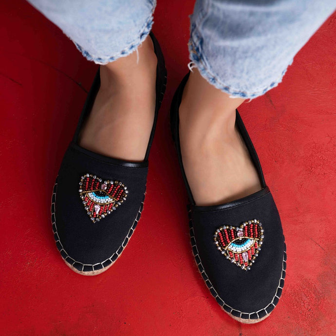 Feet of a model wearing black coloured Sweetheart Espadrilles ladies shoes with an evil eye protector design standing on a red surface
